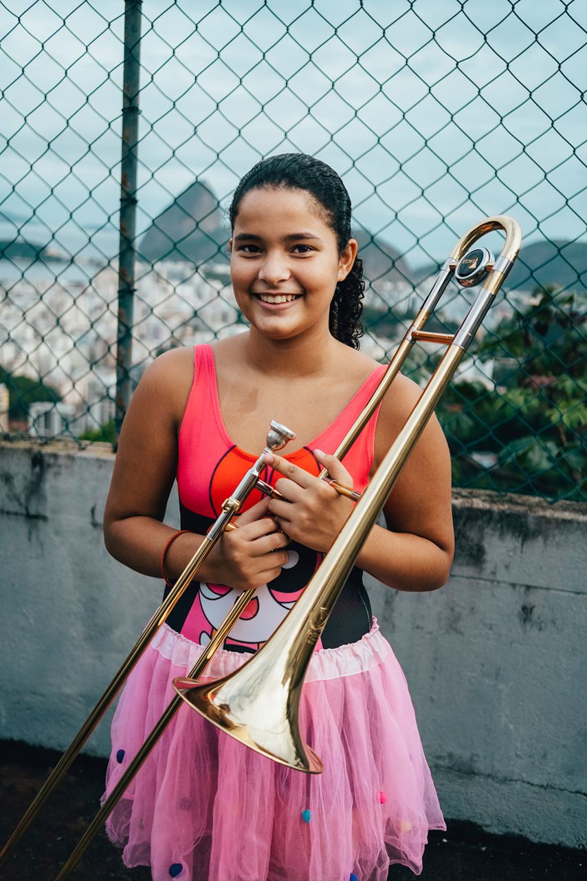 A young Favela Brass student in a pink tutu smiling and holding a trombone, with Rio de Janeiro in the background