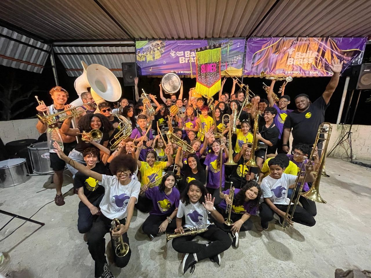 A large group of young Favela Brass musicians in yellow and purple shirts celebrating on stage with their instruments